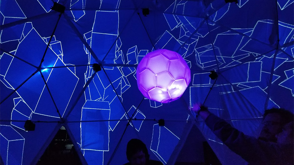 Art installation with people inside a dark room with the walls covered with abstract blue squares and a large floating pink ball in the middle