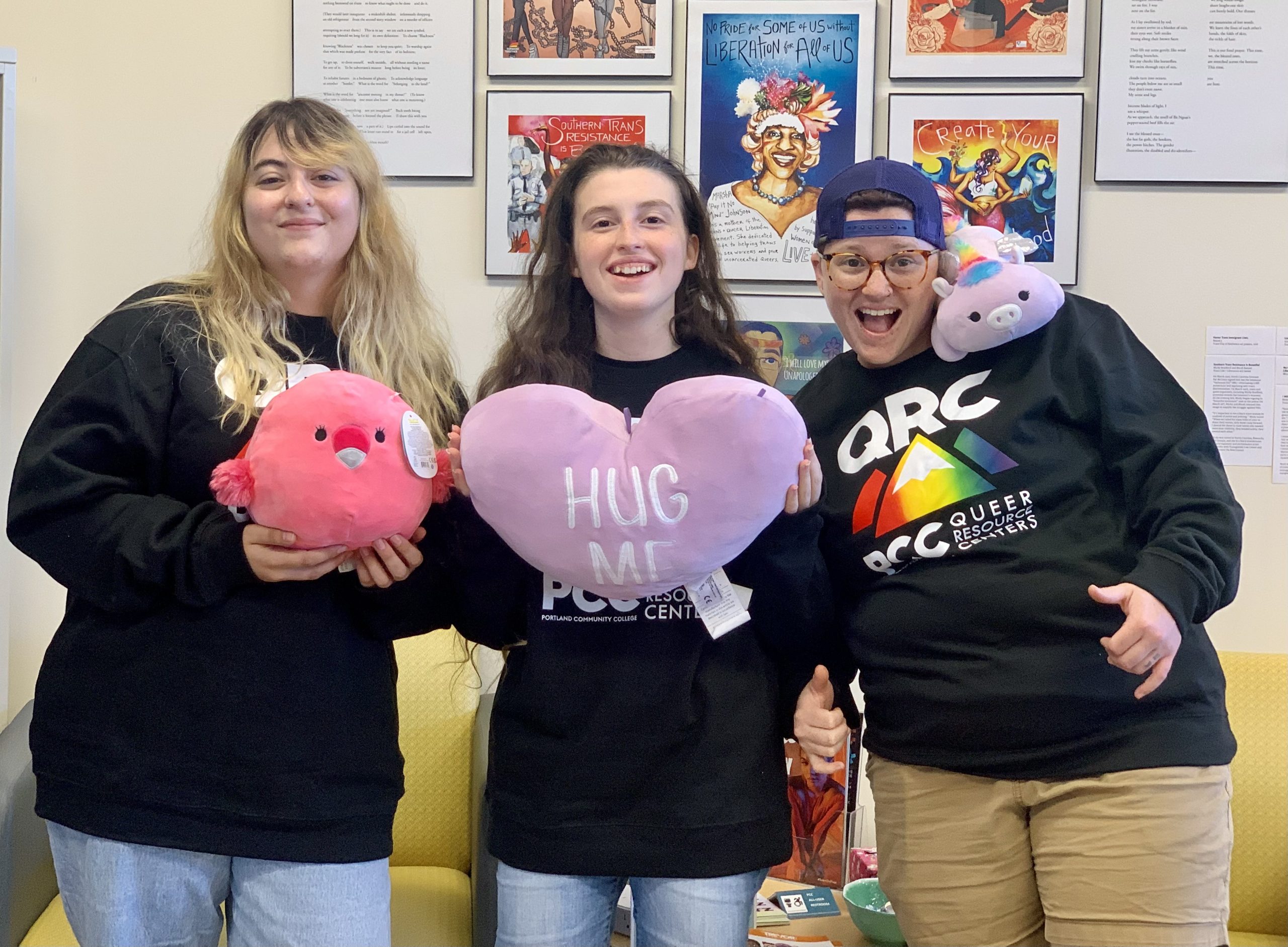 Three People are smiling and standing in front of a wall of art, all holding stuffed animals.