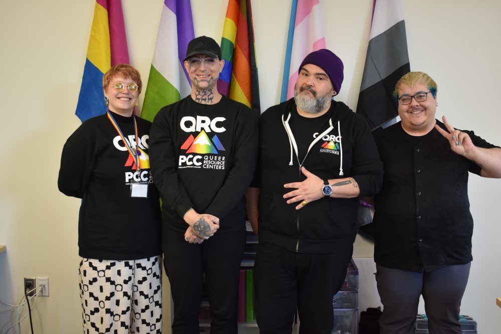 Four people in black QRC sweatshirts standing in front of rainbow flags.