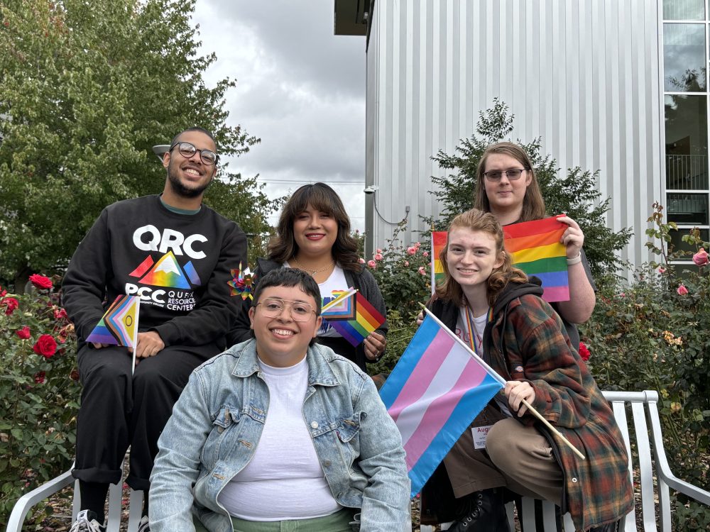 four students in QRC sweatshirts hold rainbow and trans flags behind their coordinator who is seated on a bench wearing a jean jacket.