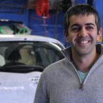 Business owner smiles while standing in the lobby of his business, with two men working behind counter in the background