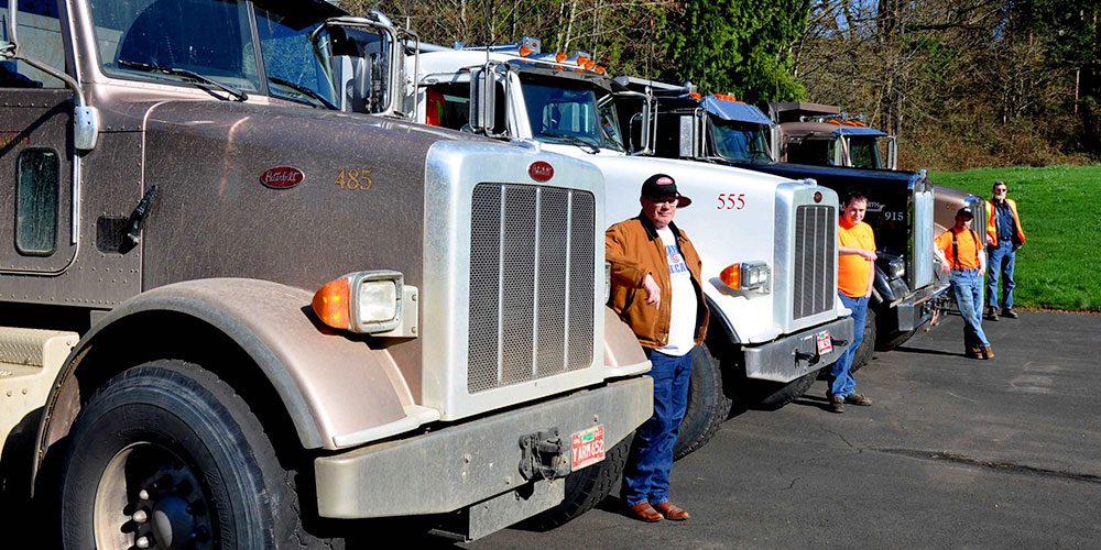Line of 4 semi trucks recedes into distance, with Bud Construction workers standing in front of each truck