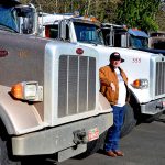 Line of 4 semi trucks recedes into distance, with Bud Construction workers standing in front of each truck