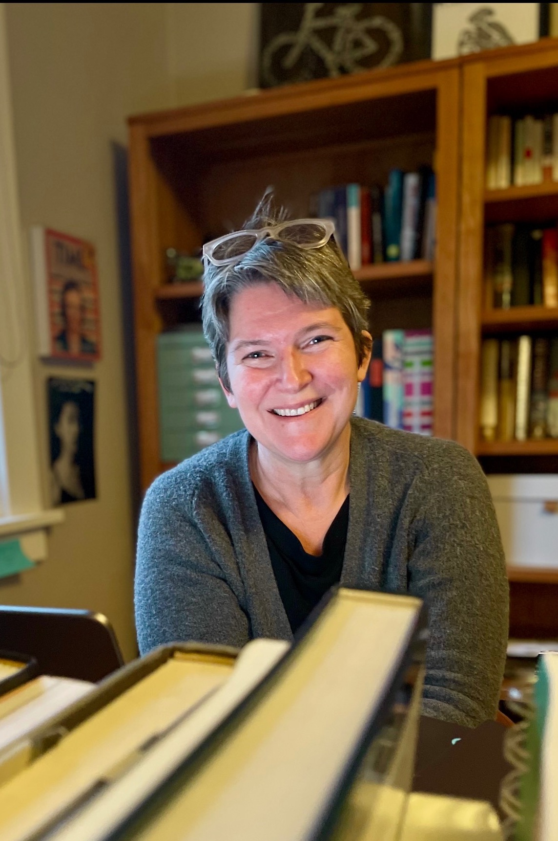 woman with short brown and gray hair and reading glasses on her head smiling from behind a desk piled with books