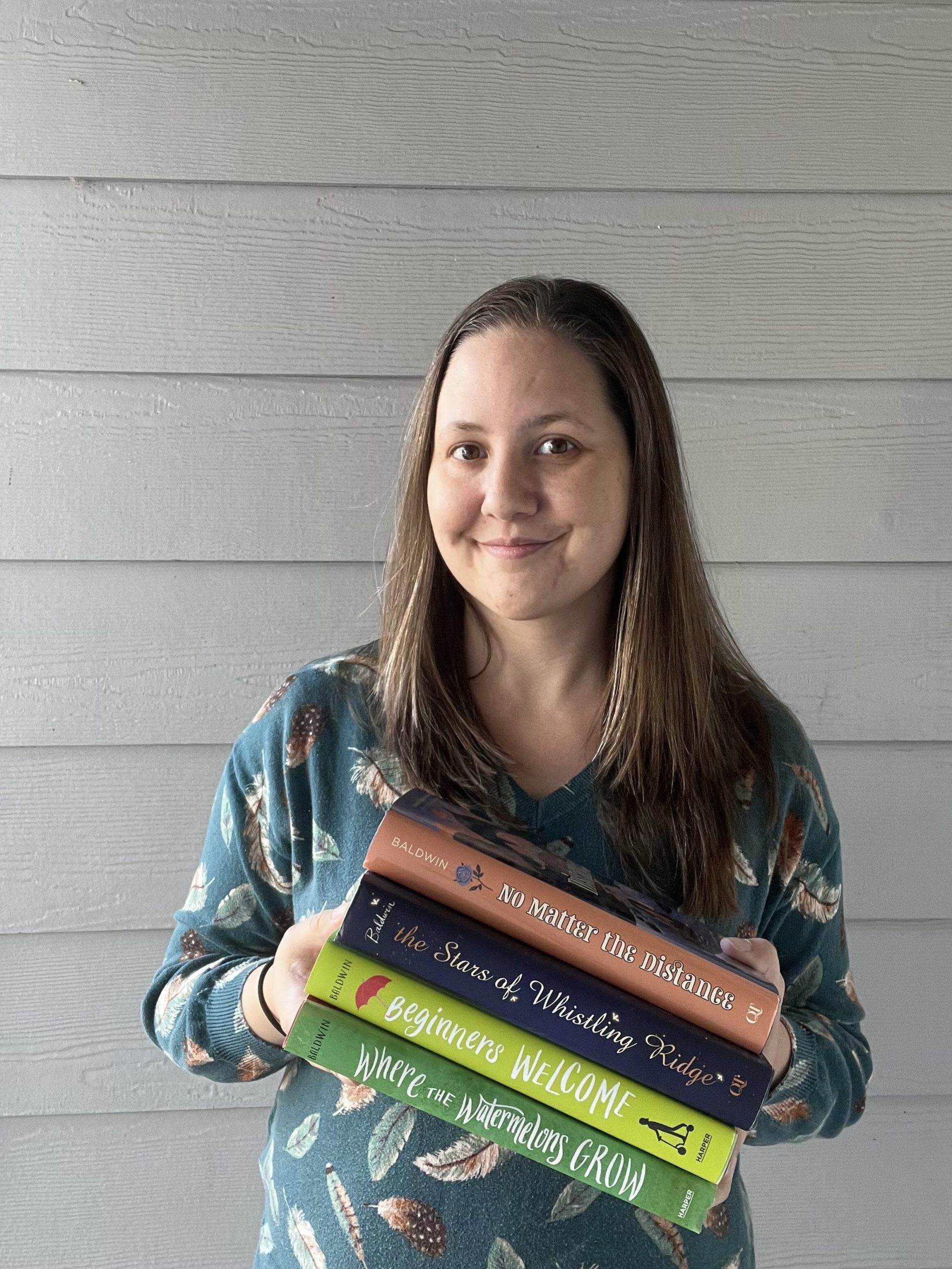 Cindy, a white woman with brown hair wearing a green sweater with a feather print design, holds four books: NO MATTER THE DISTANCE, THE STARS OF WHISTLING RIDGE, BEGINNERS WELCOME, and WHERE THE WATERMELONS GROW.