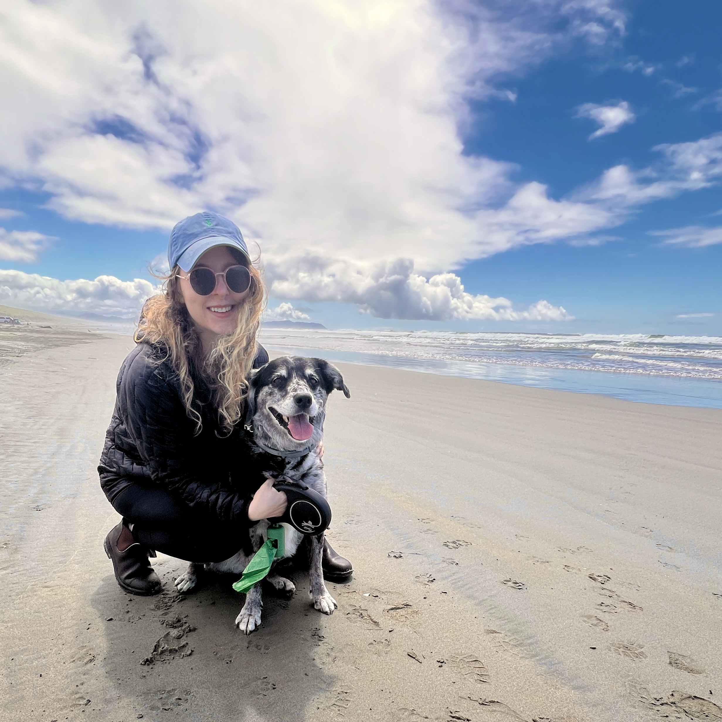 A woman wearing round sunglasses, a light blue baseball cap, and a black jacket crouches on the wet sand of a wide beach next to her speckled black-and-grey dog. The dog is sitting upright, looking happy with its tongue out, attached to a retractable leash held by the woman. In the background, ocean waves crash along the shoreline beneath a bright blue sky filled with puffy white clouds.