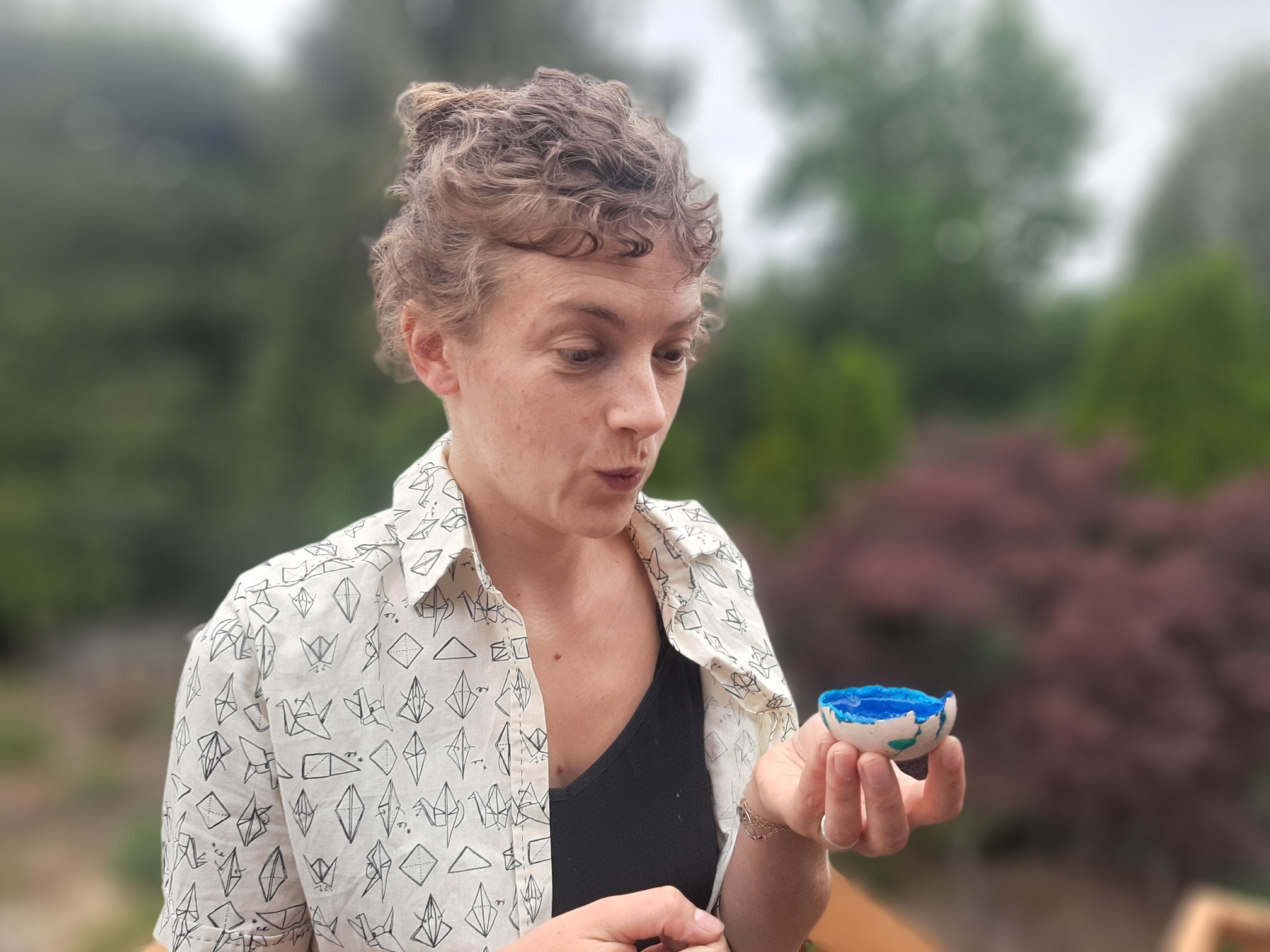 Your course instructor, a white woman in her 30s with curly hair wearing a shirt with an origami print, is looking surprised by an eggshell with something crystallised and blue in it.