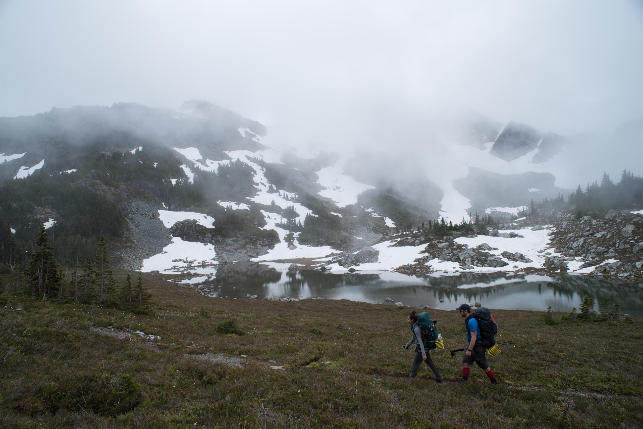 Two people backpacking through misty mountains, near a small lake. Patchy snow behind the hikers.