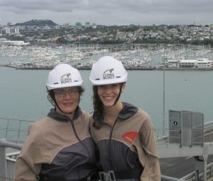 Two women in safety gear on the Auckland bridge with view of the marina and city.
