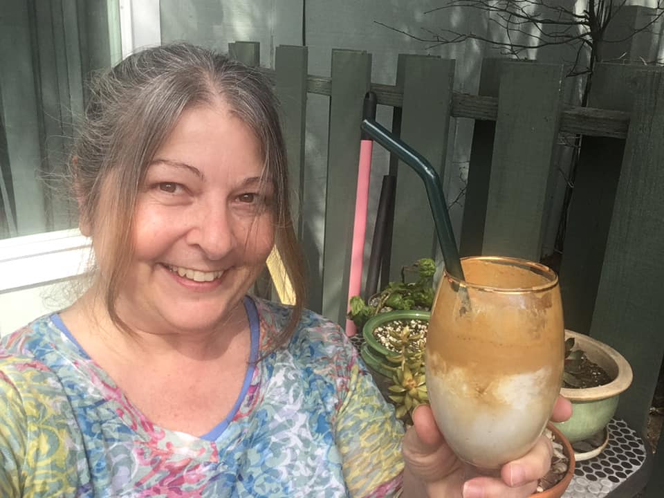 Mary Anne sitting outside on the patio while smiling and holding up her homemade dalgona coffee drink.