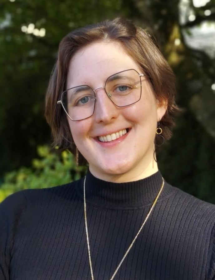 Miranda smiling in front of a lush green background. She has short brown hair and light skin, and wears thin framed glasses, a black knit shirt, and a gold necklace.