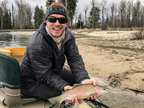PCC fly fishing instructor Nick Parish holds a rainbow trout while sat in the front seat of a drift boat on a river somewhere in Montana.