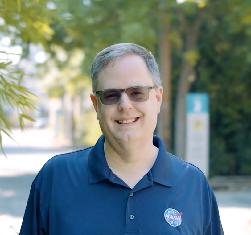 A picture of myself wearing a blue NASA polo shirt outside on the Cascade Campus with trees behind me