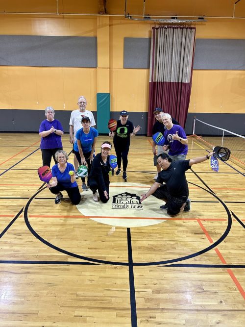 Pickleball players holding paddles and standing or kneeling around a logo on a gym floor that says "Friendly House".