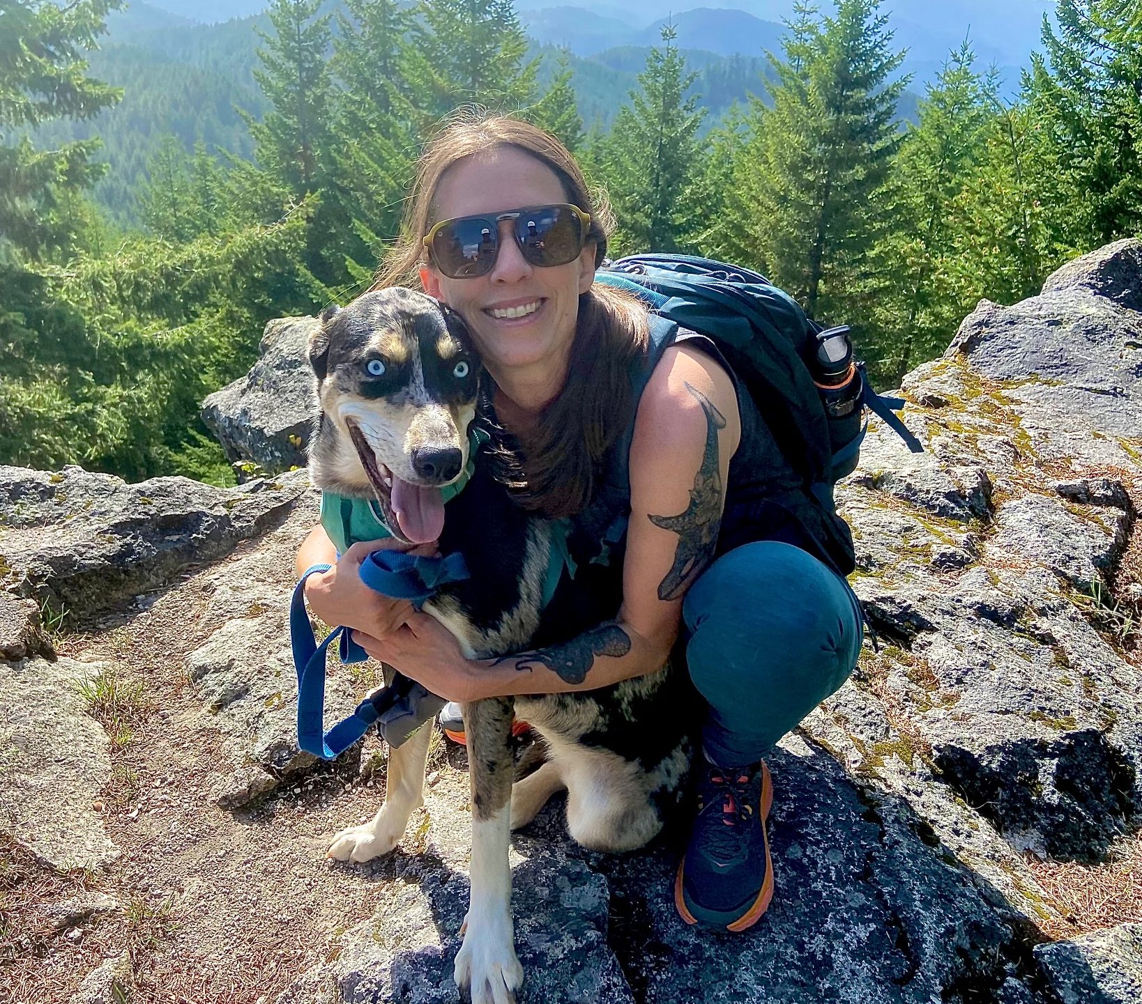 A photo of Taryn wearing sunglasses and a backpack. She is crouched down hugging her dog, Piper, who is a mixed breed. You can see trees and mountain in the background.