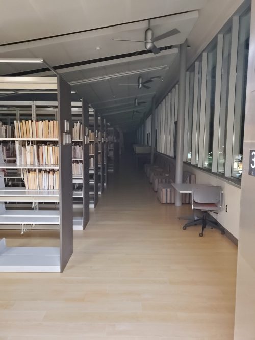 The third floor of Southeast Campus Naito Family library, viewed from the elevator on a dark evening. The stacks and study tables run along the sides of the viewers perception until they hit a point of total darkness.
