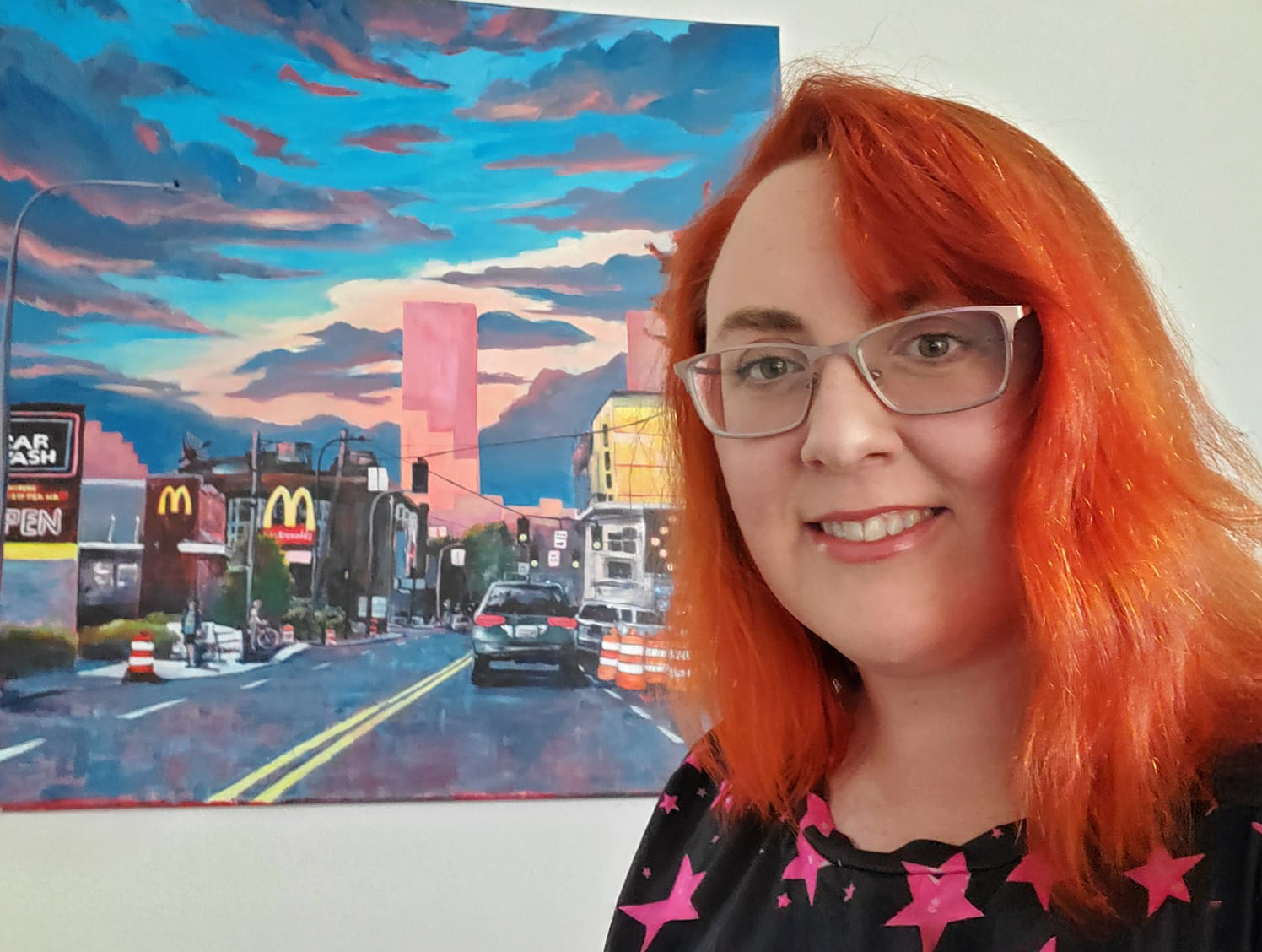 A picture of a red haired, smiling woman with glasses in front of a cityscape painting