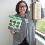 smiling woman raises compost bin and compost instructions poster