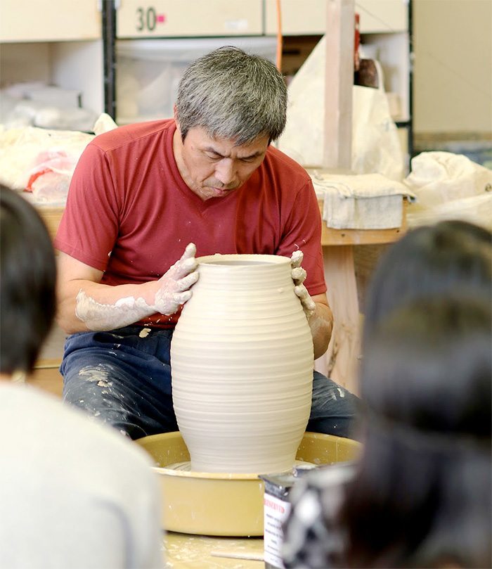 Photo of a potter - two students in front of him are blurred out, as is the background