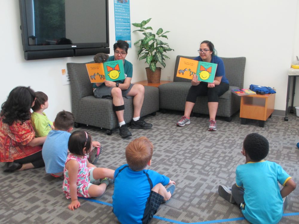 Rut Martínez-Alicea, coordinator for the Multicultural Center (left), listens along with the children from the YMCA childcare center as Farnoosh Khodayar (center) and Elmira Fathe Azam read aloud from a book on diversity. Between six and 10 children attend the story time every Tuesday morning. 