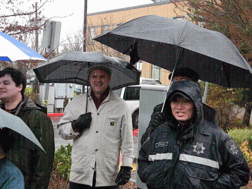 Campus and community members hover on the edges of the event tent with the protection of umbrellas. 